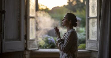 Woman in a light-colored outfit exhaling smoke while standing by an open window with sunlight filtering through.