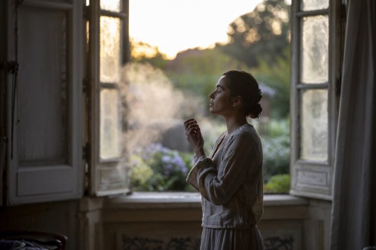 Woman in a light-colored outfit exhaling smoke while standing by an open window with sunlight filtering through.