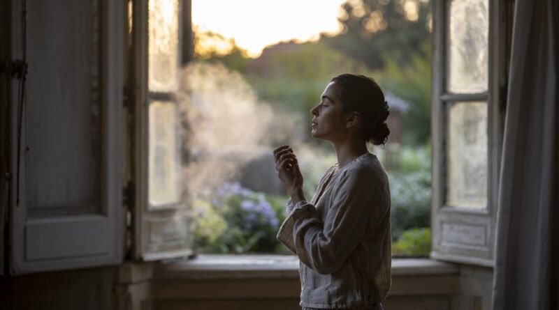 Woman in a light-colored outfit exhaling smoke while standing by an open window with sunlight filtering through.