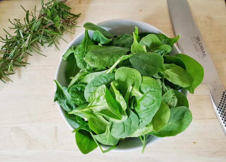 Fresh spinach leaves arranged in a white bowl, with a sprig of rosemary and a kitchen knife nearby.