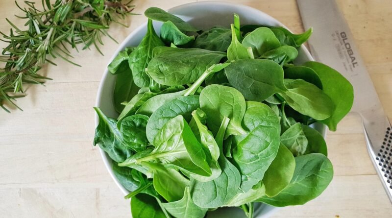 Fresh spinach leaves arranged in a white bowl, with a sprig of rosemary and a kitchen knife nearby.