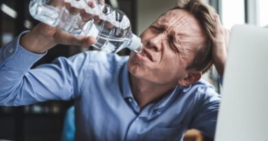 Man in a blue shirt drinking water from a plastic bottle while showing signs of fatigue at an office desk with papers and a laptop.